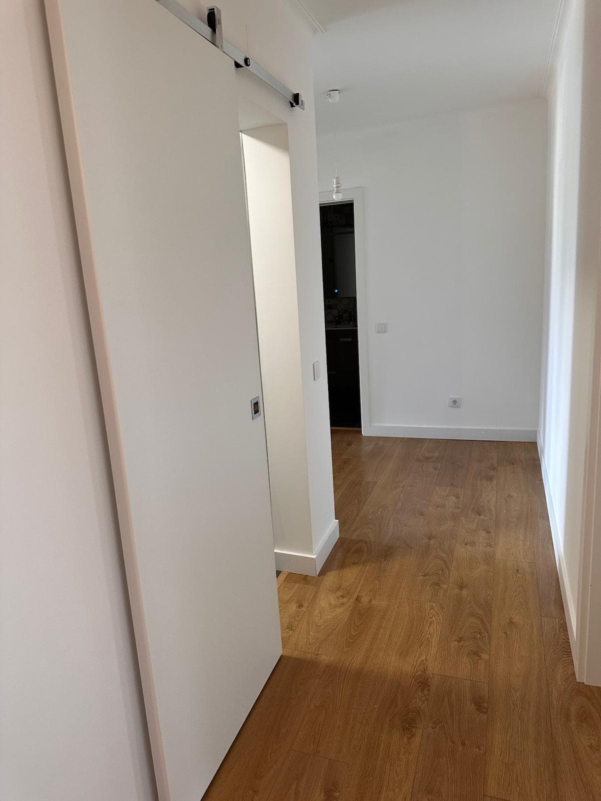 Corredor renovado de apartamento com porta de correr em metal, paredes brancas e soalho de madeira clara.